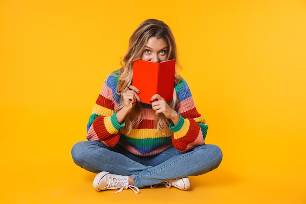 A girl holds a book while sitting on the floor, against a yellow wall